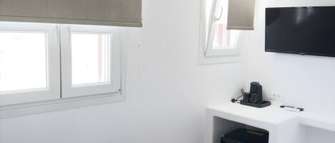 A modern bedroom with a bed, a desk with a computer and a coffee maker, a TV on the wall, and a window with a blind.