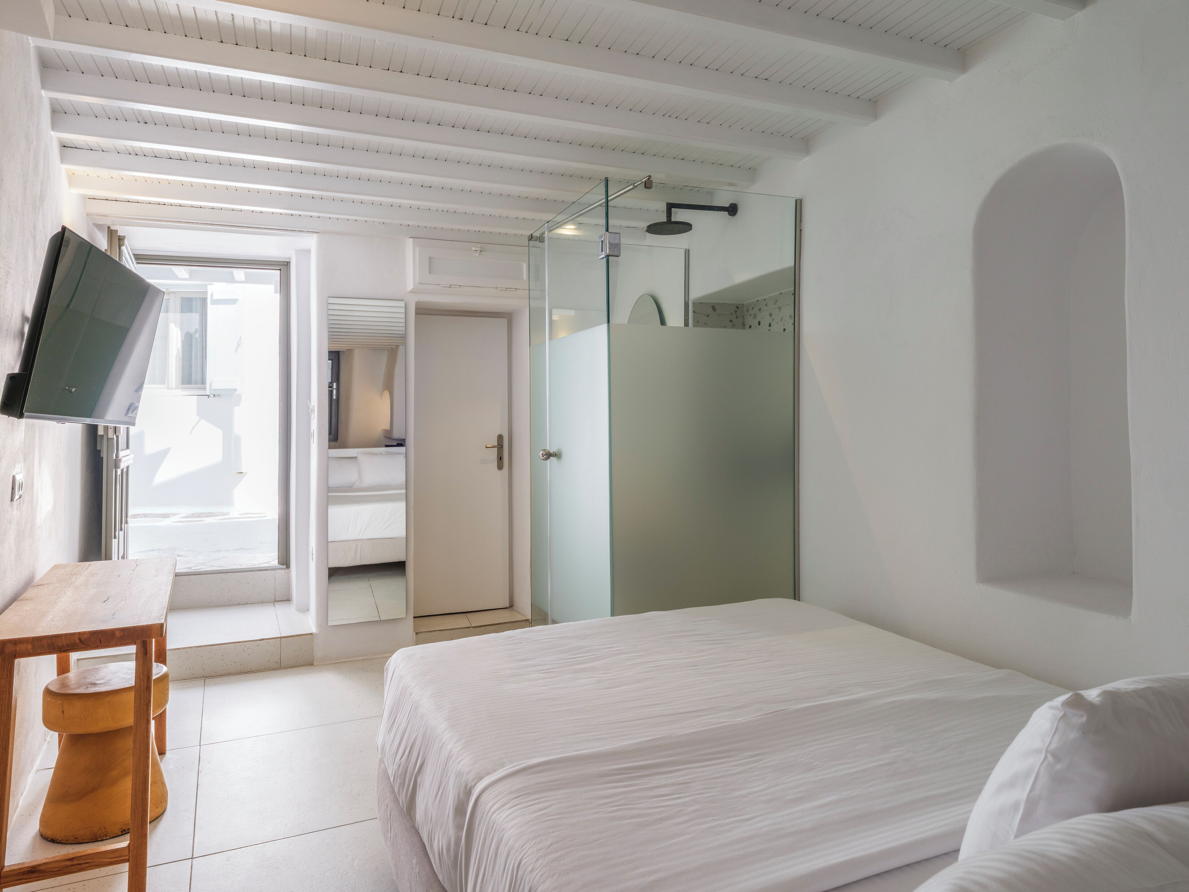 Una habitación de hotel moderna con una cama, una mesa de madera, un televisor montado en la pared y un rincón de ducha.
