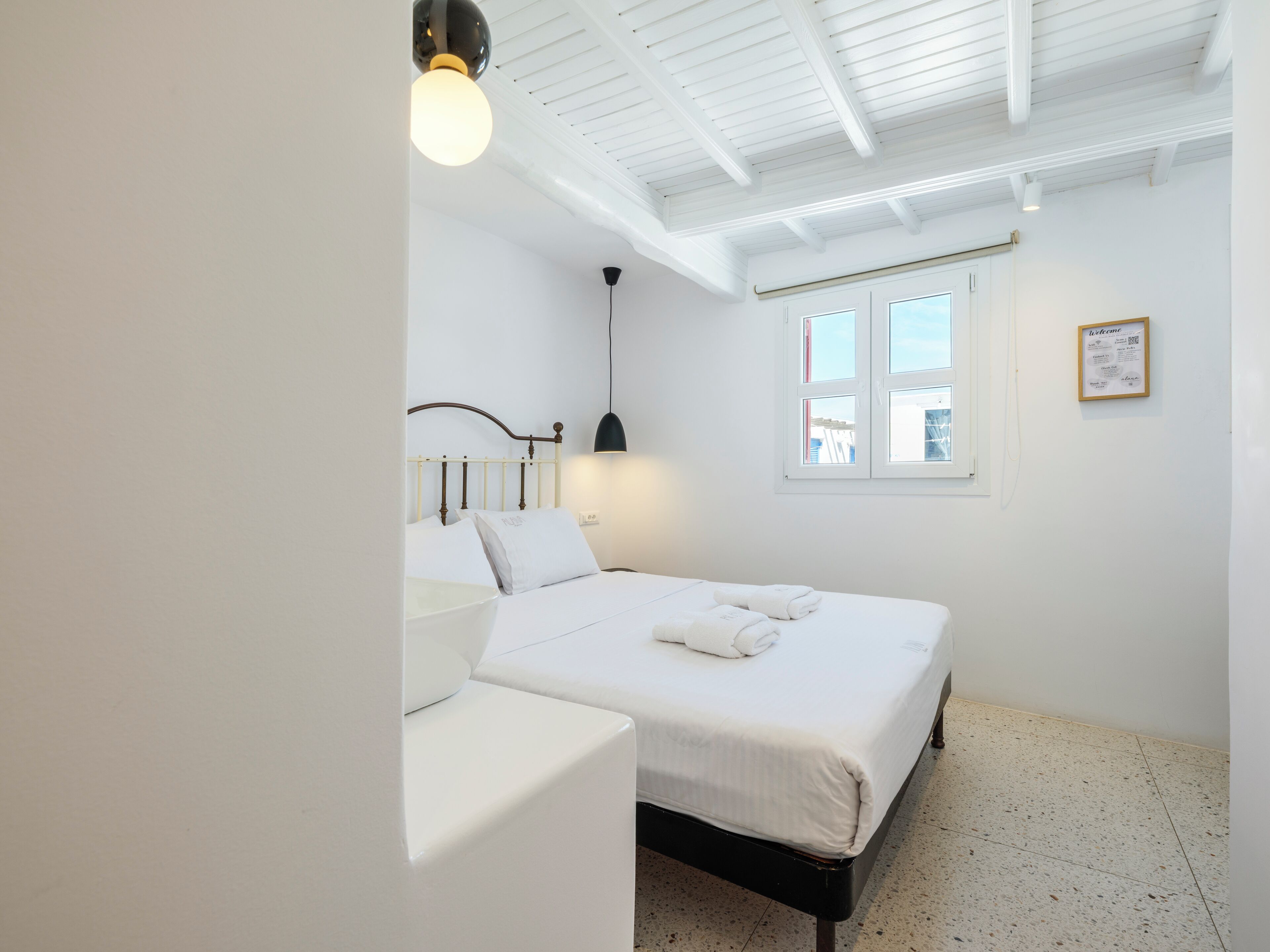 Una habitación limpia y blanca con una cama, una mesita de noche, una lámpara y una ventana.