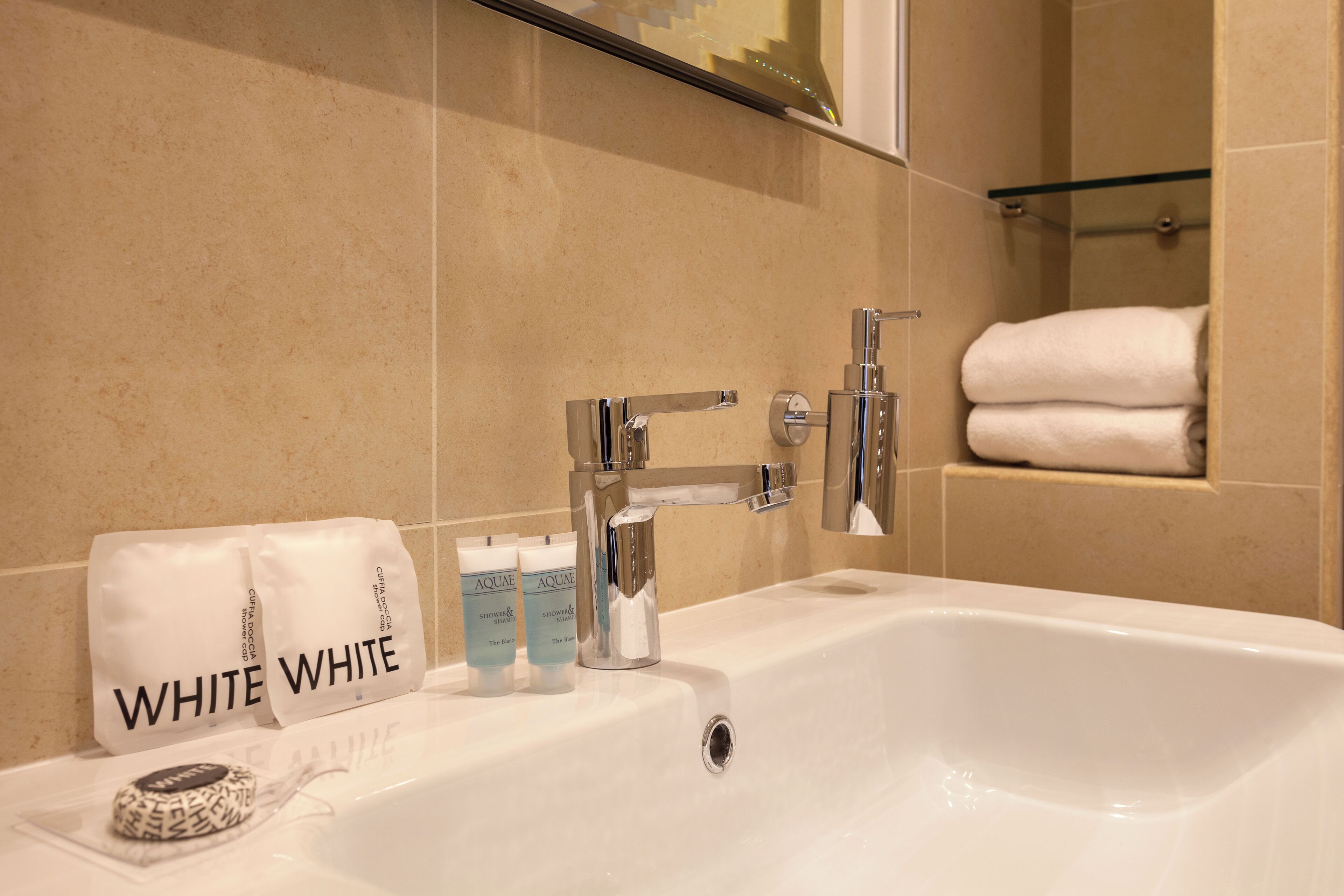 Un baño con un lavamanos blanco, dos botellas de champú 'AQUA WHITE', una toalla y un estante de vidrio.