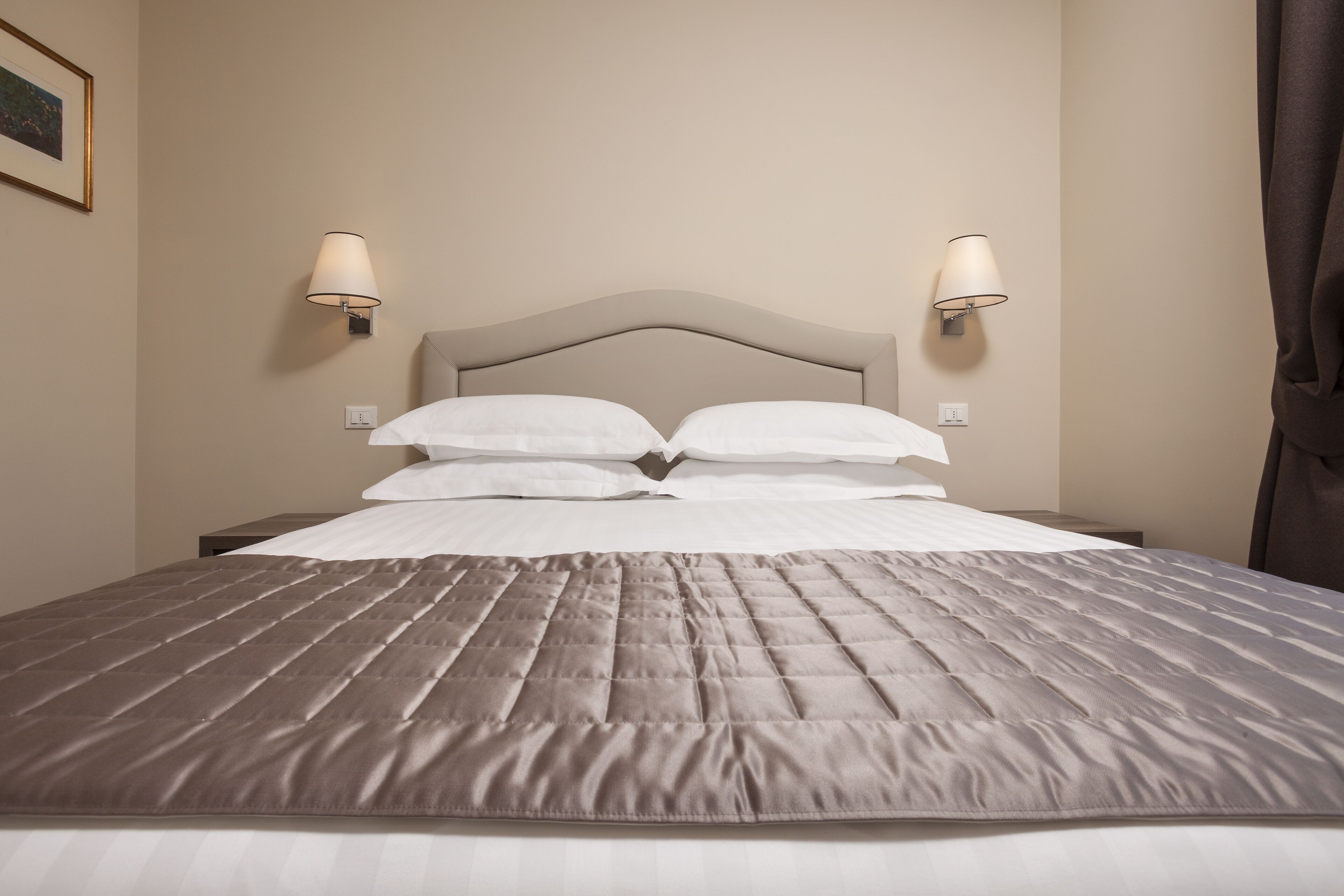 Una cama bien hecha con cabecera, dos lámparas fijadas a la pared y un cuadro enmarcado en la pared.