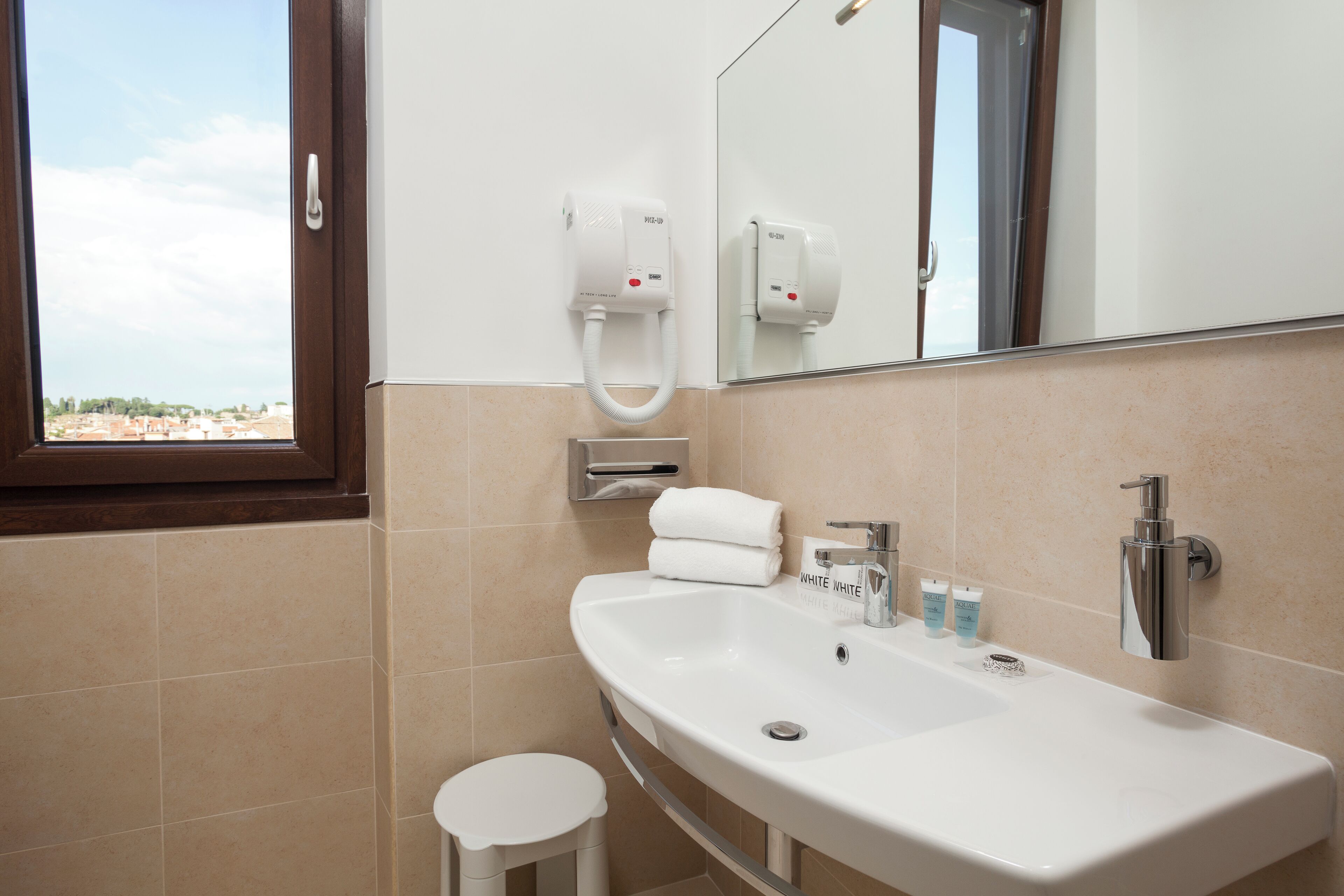 Un baño con un lavamanos blanco, un espejo, un dispensador de toallas y una ventana con vista a edificios.