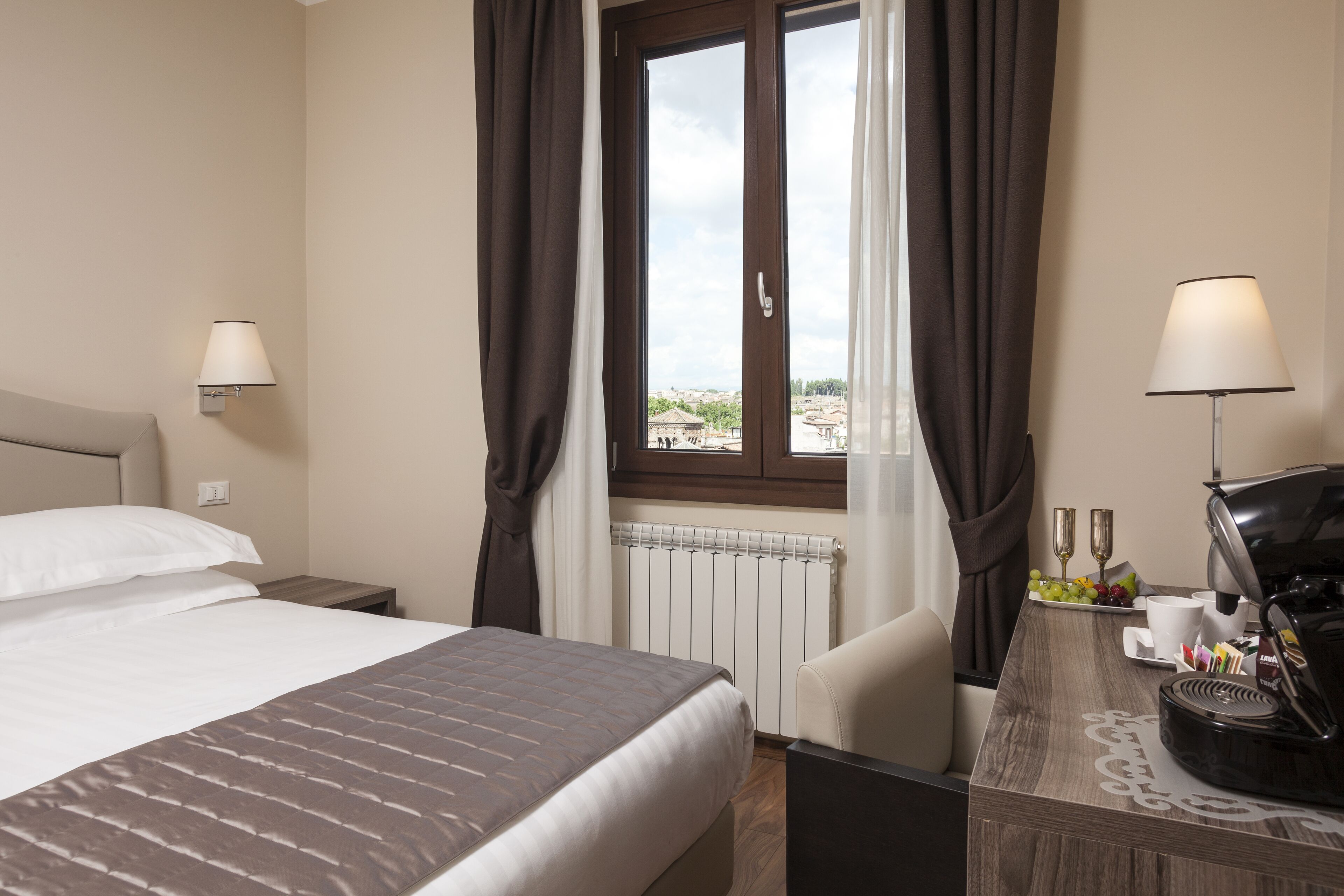 Une chambre d’hôtel comprenant un lit, un bureau avec une machine à café, deux lampes fixées au mur, une fenêtre avec des rideaux et une vue sur les bâtiments environnants.