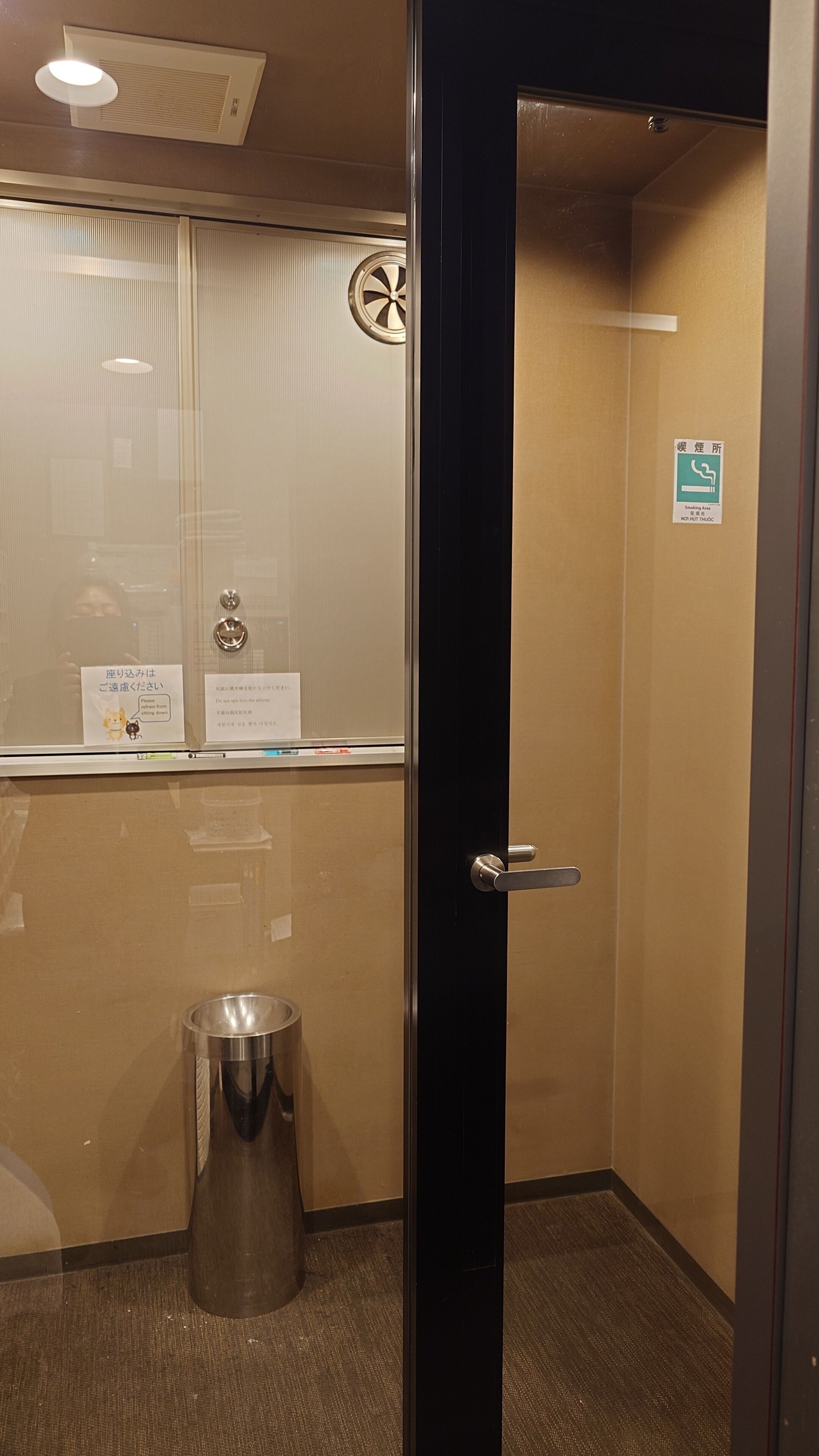 A small, enclosed restroom with a trash bin, a hand sanitizer dispenser, and a sign indicating a smoking area.
