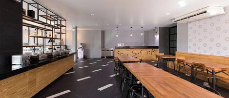 A modern restaurant with a black and white color scheme, featuring a long wooden counter, multiple seating areas, and a kitchen in the background.