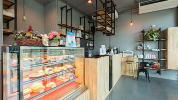 Una panadería con un expositor de vidrio lleno de pasteles y un mostrador con productos horneados. Se ven estantes con flores y un cartel.