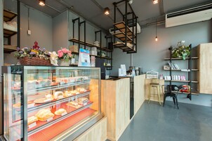 Una panadería con un expositor de vidrio lleno de pasteles y un mostrador con productos horneados. Se ven estantes con flores y un cartel.