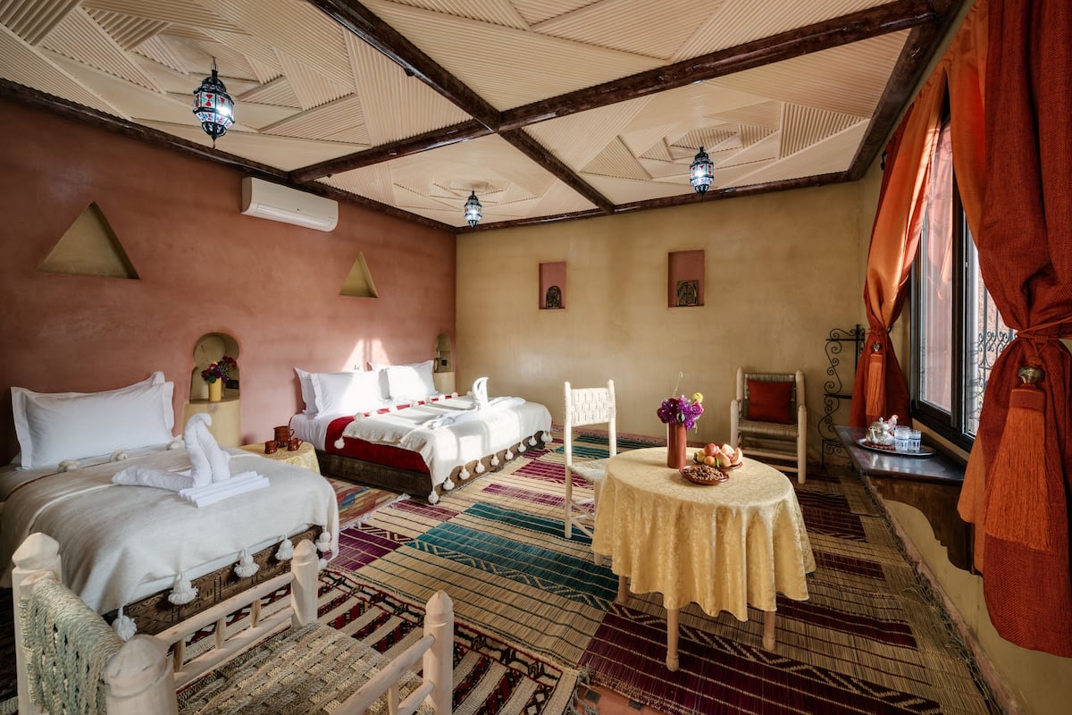 Une chambre d’hôtel avec deux lits, une table à manger, une chaise, une fenêtre avec des rideaux et un ventilateur de plafond.