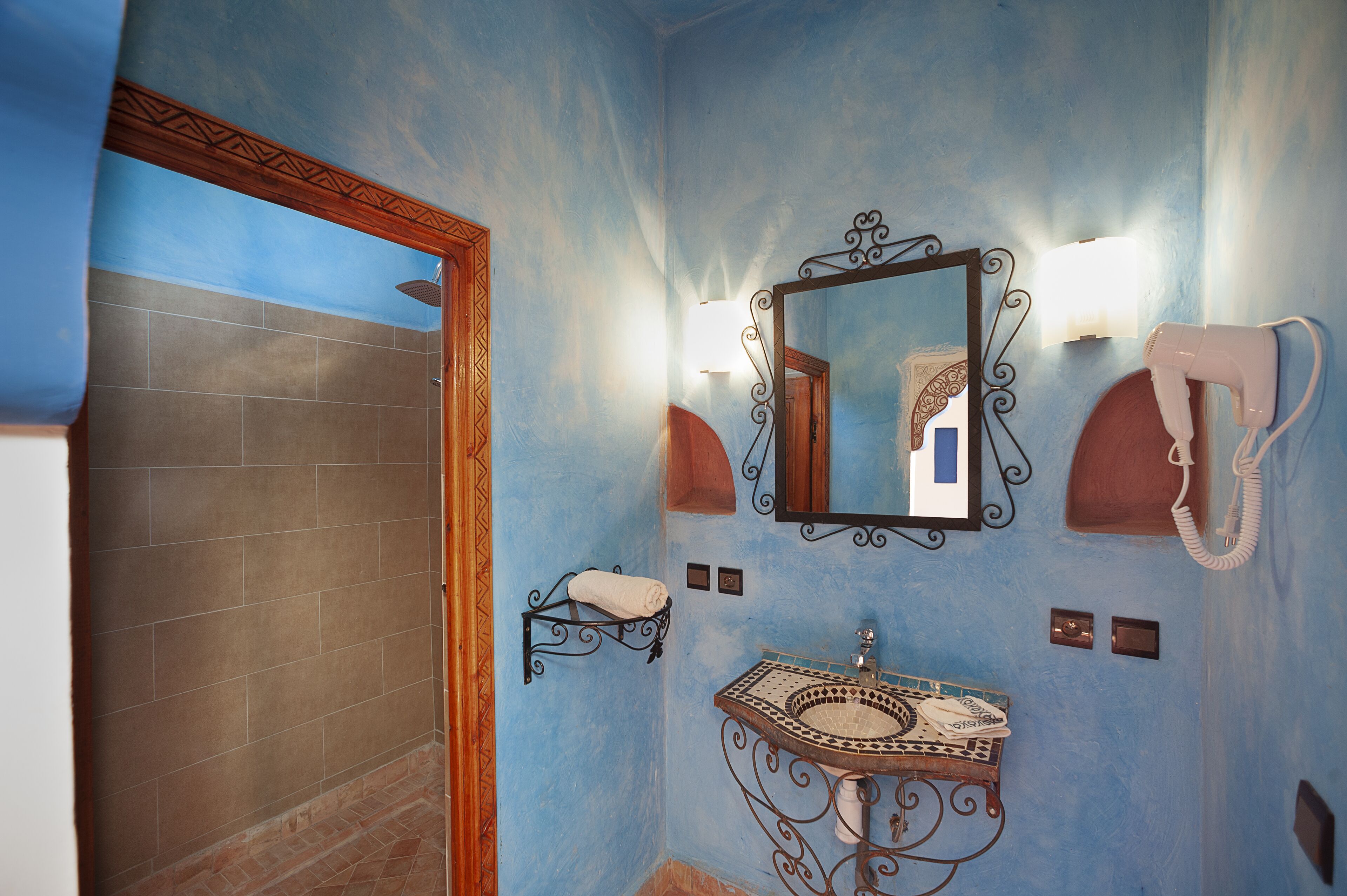A bathroom with a blue wall, a wooden door, a sink with a decorative basin, a mirror with a decorative frame, a towel rack, and a hairdryer.