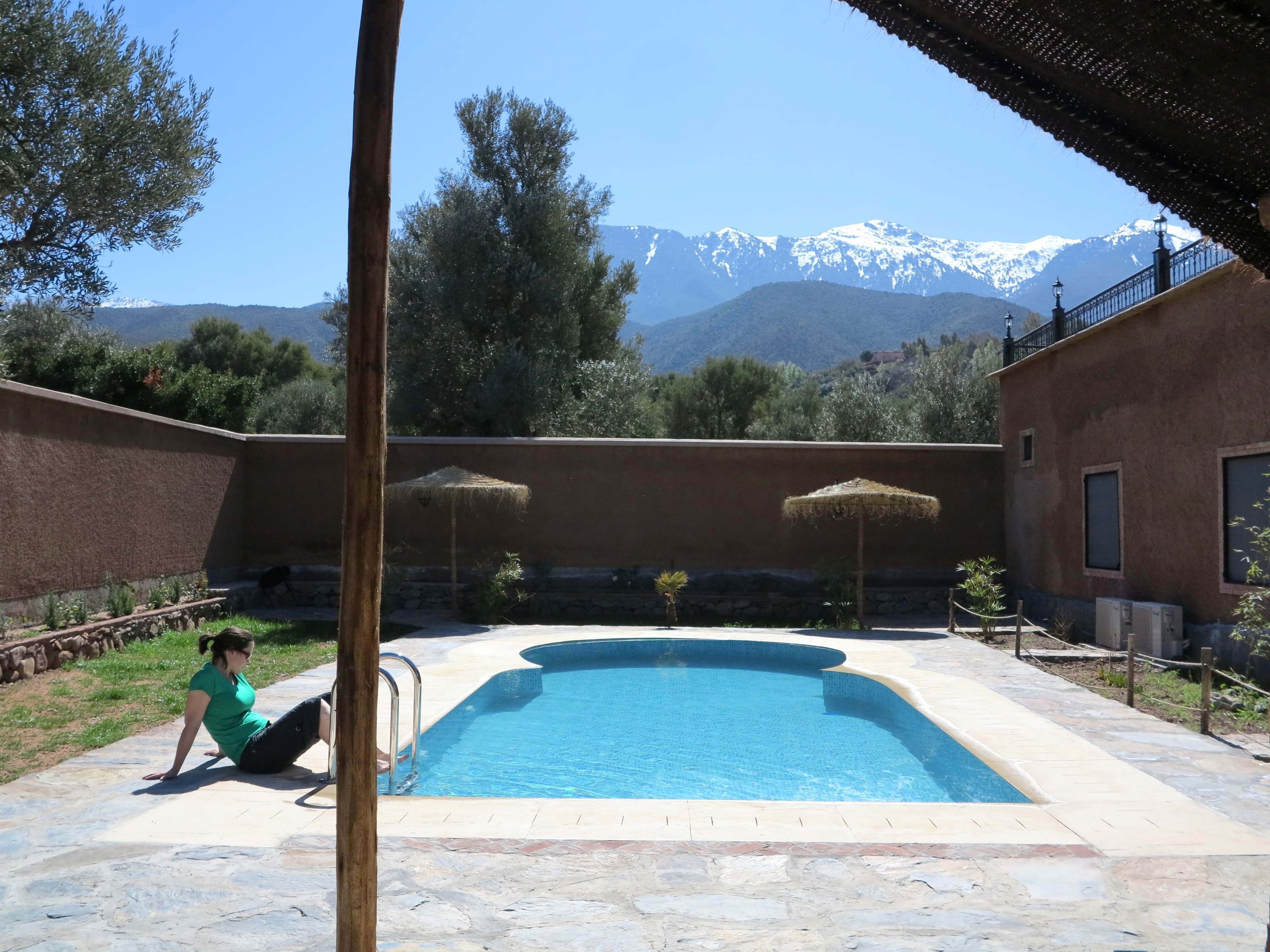 A swimming pool with a stone deck, a person sitting by the pool, and mountains in the background.