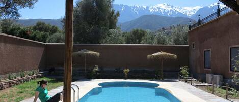 Ein Schwimmbad mit einer Steindeck, eine Person sitzt am Pool, im Hintergrund Berge.