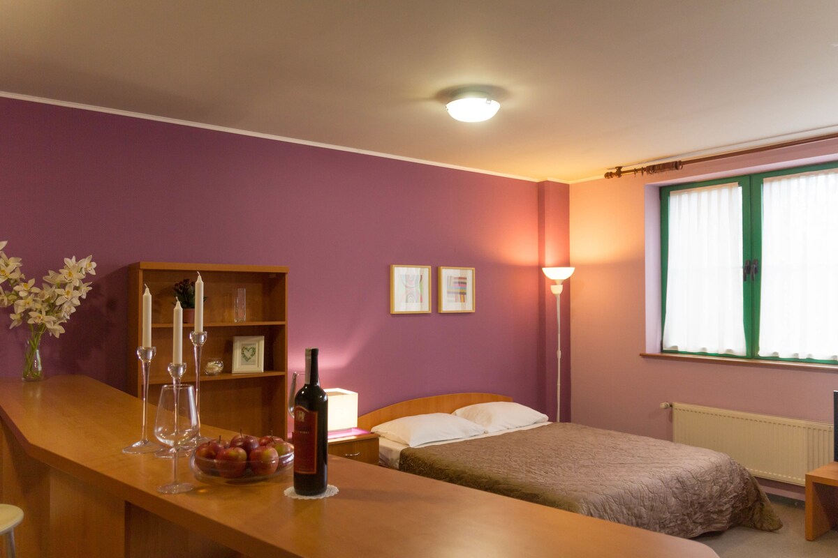 Habitación de hotel con un escritorio de madera, una cama con cubrecama color beige, una mesita de noche con lámpara y una ventana con cortinas verdes.