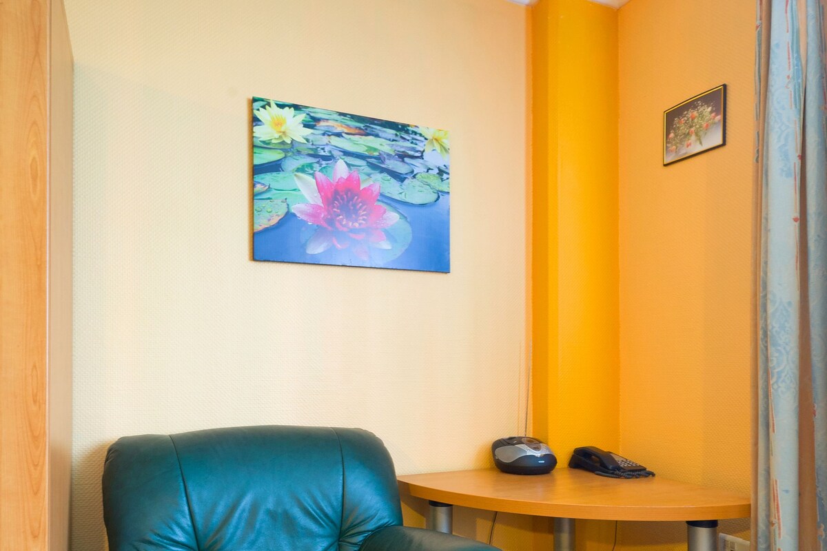 Una habitación con un sillón de cuero verde, una mesita de madera, un cuadro de flores y una cortina.