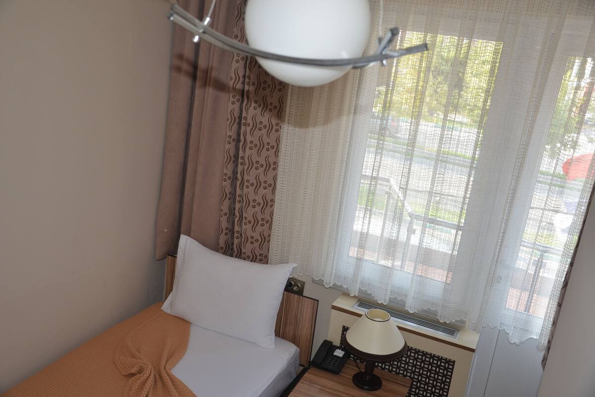 A bedroom with a bed, a bedside table with a lamp, a window with curtains, and a wall-mounted air conditioning unit.