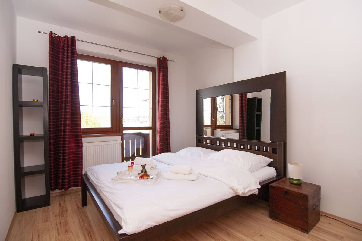 A bedroom with a wooden bed, a nightstand, a dresser, a window with curtains, and a shelf unit.