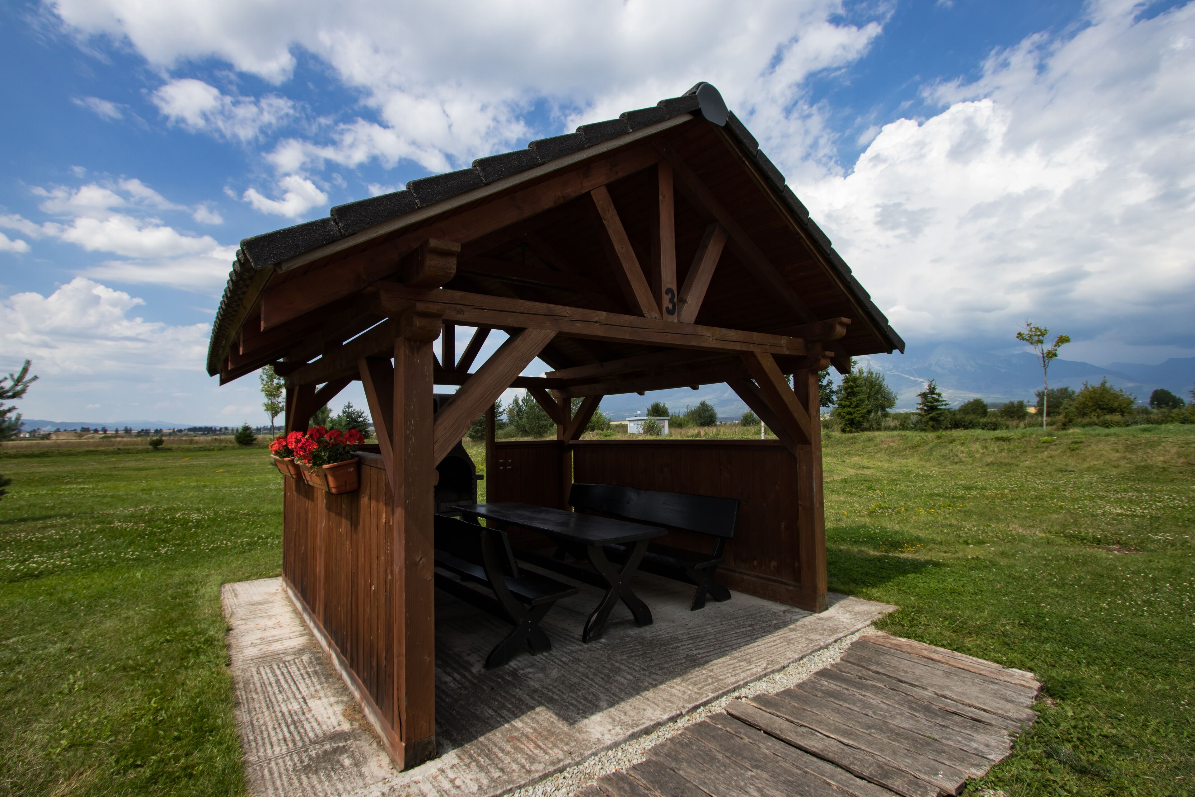 A wooden gazebo with a bench and picnic table, surrounded by a grassy field and a clear sky.