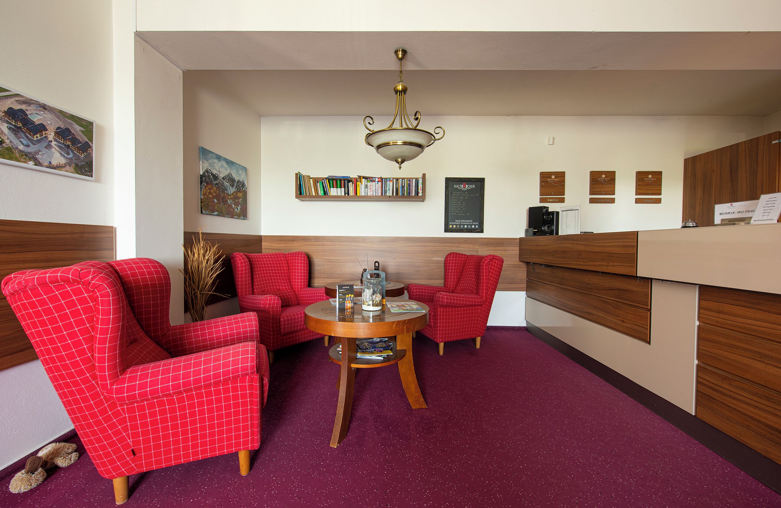 Un vestíbulo de hotel con sillas de cuadros rojos, una mesa redonda de madera, una araña, una estantería y un mostrador de recepción.
