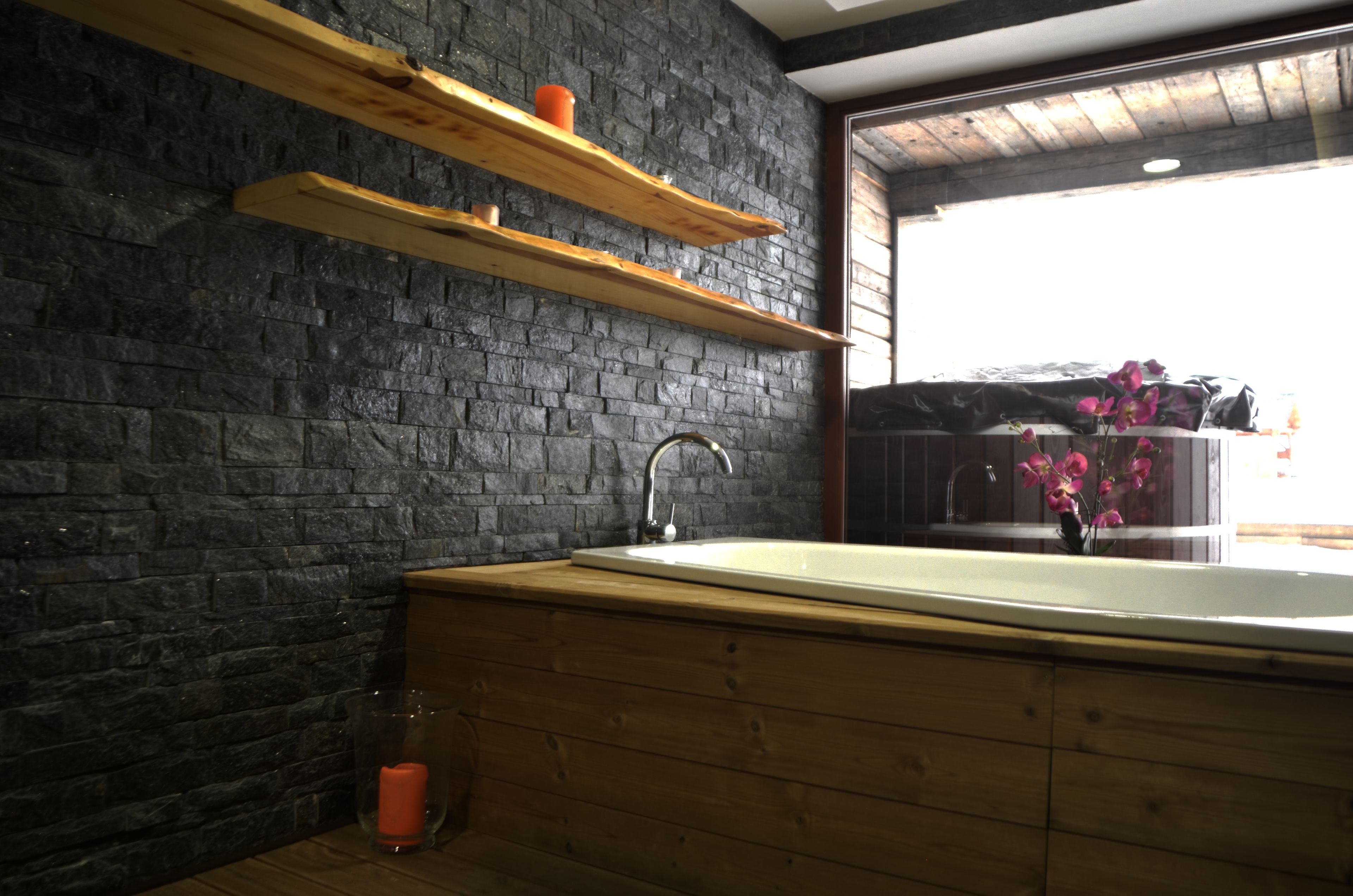 A modern bathroom with a stone wall, wooden shelves, a large bathtub, and a window with flowers.