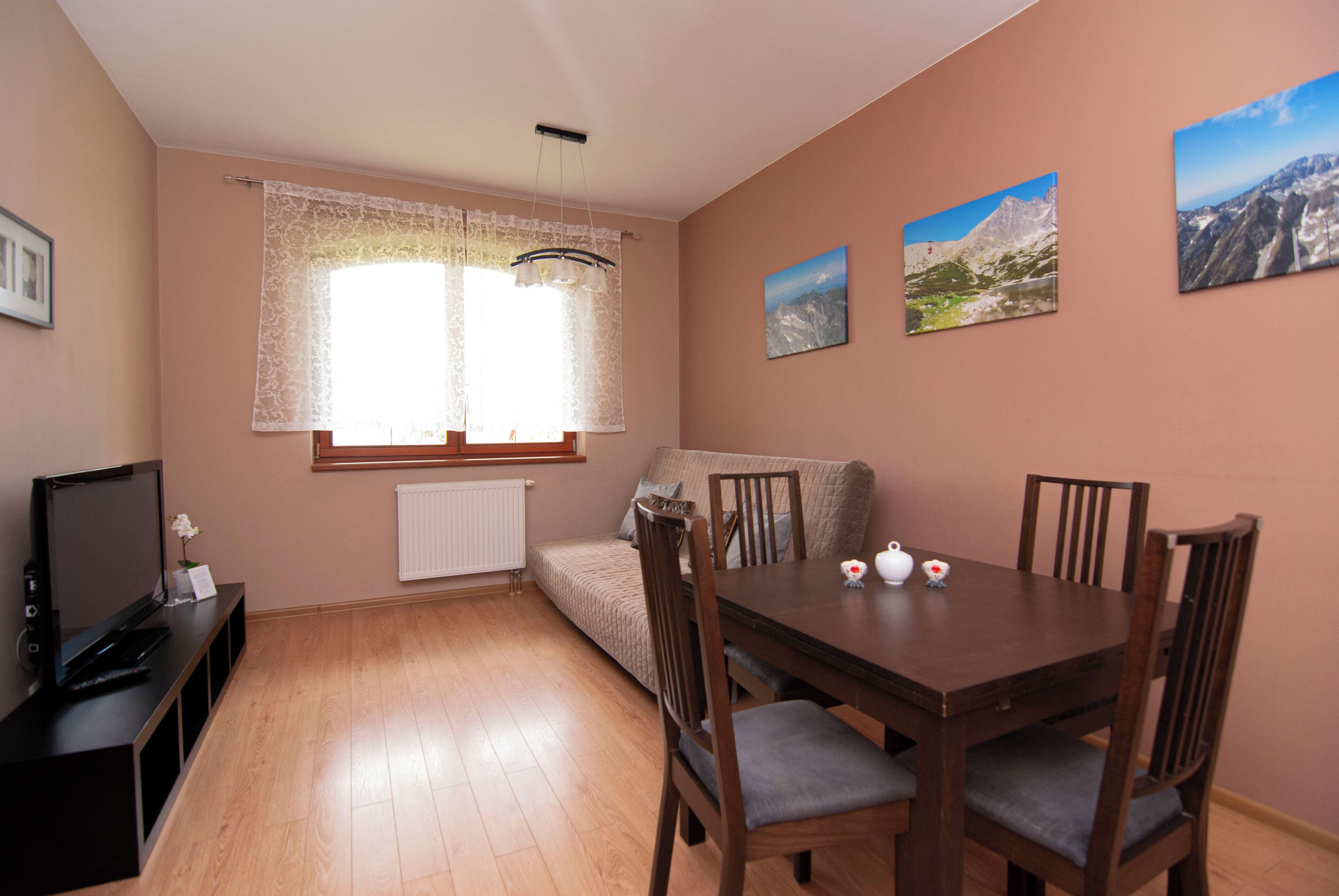 Ein Wohnzimmer mit Fernseher, Sofa, Esstisch, Stühlen und zwei Bildern an der Wand.
