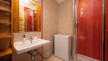 Une salle de bain avec un lavabo blanc, des étagères en bois, un miroir et une cabine de douche carrelée de carreaux rouges.
