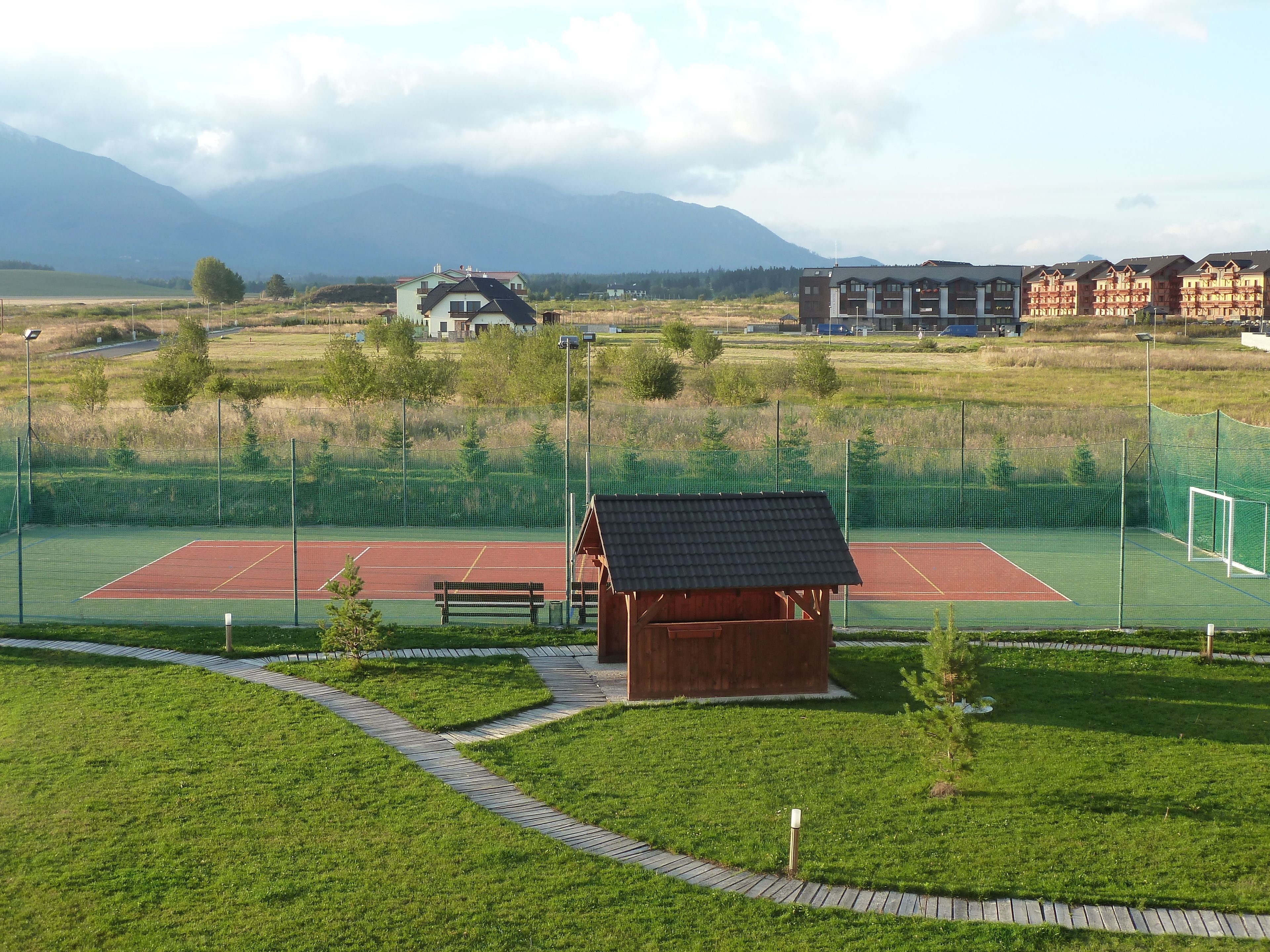 En tennisbane med en lille trækonstruktion, omgivet af et grønt hegn og en sti.