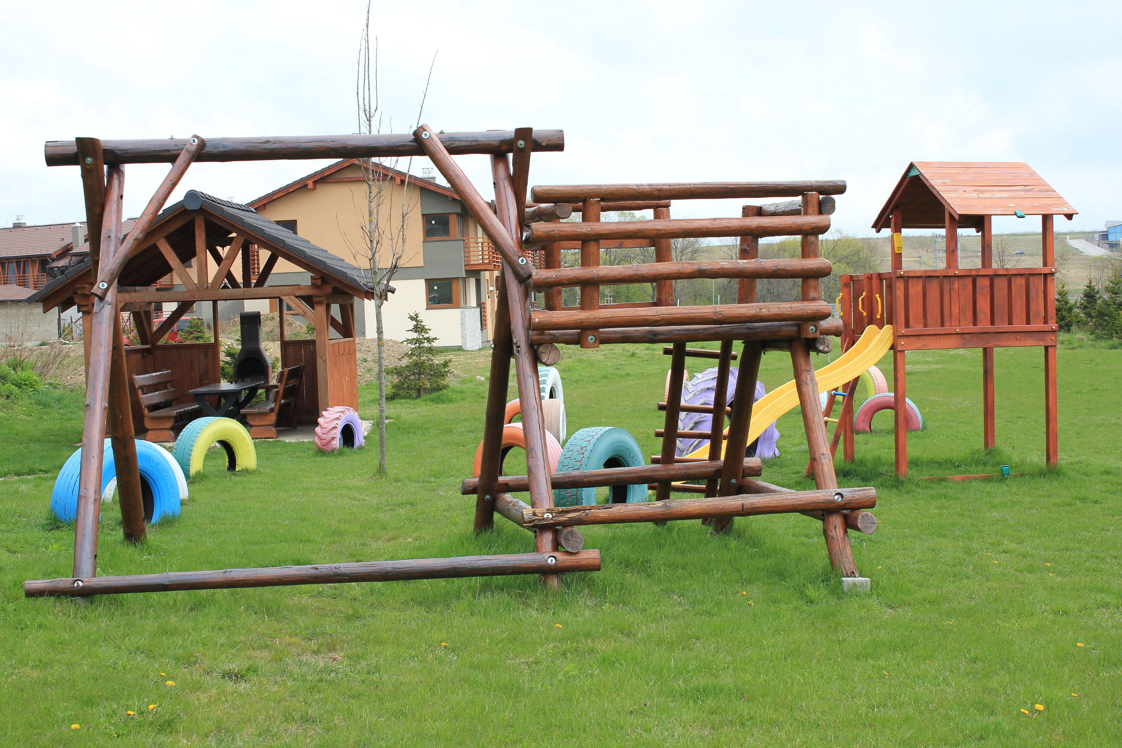 A wooden play structure with a slide and climbing elements, surrounded by grass and a building in the background.