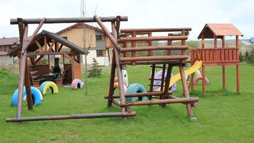A wooden play structure with a slide and climbing elements, surrounded by grass and a building in the background.
