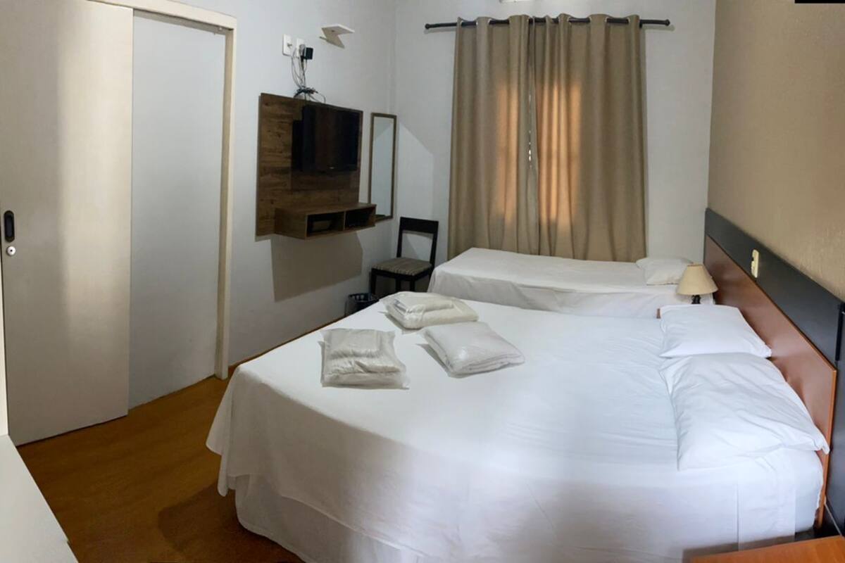 Habitación de hotel con dos camas, un televisor montado en un panel de madera, una silla y una ventana con cortinas.