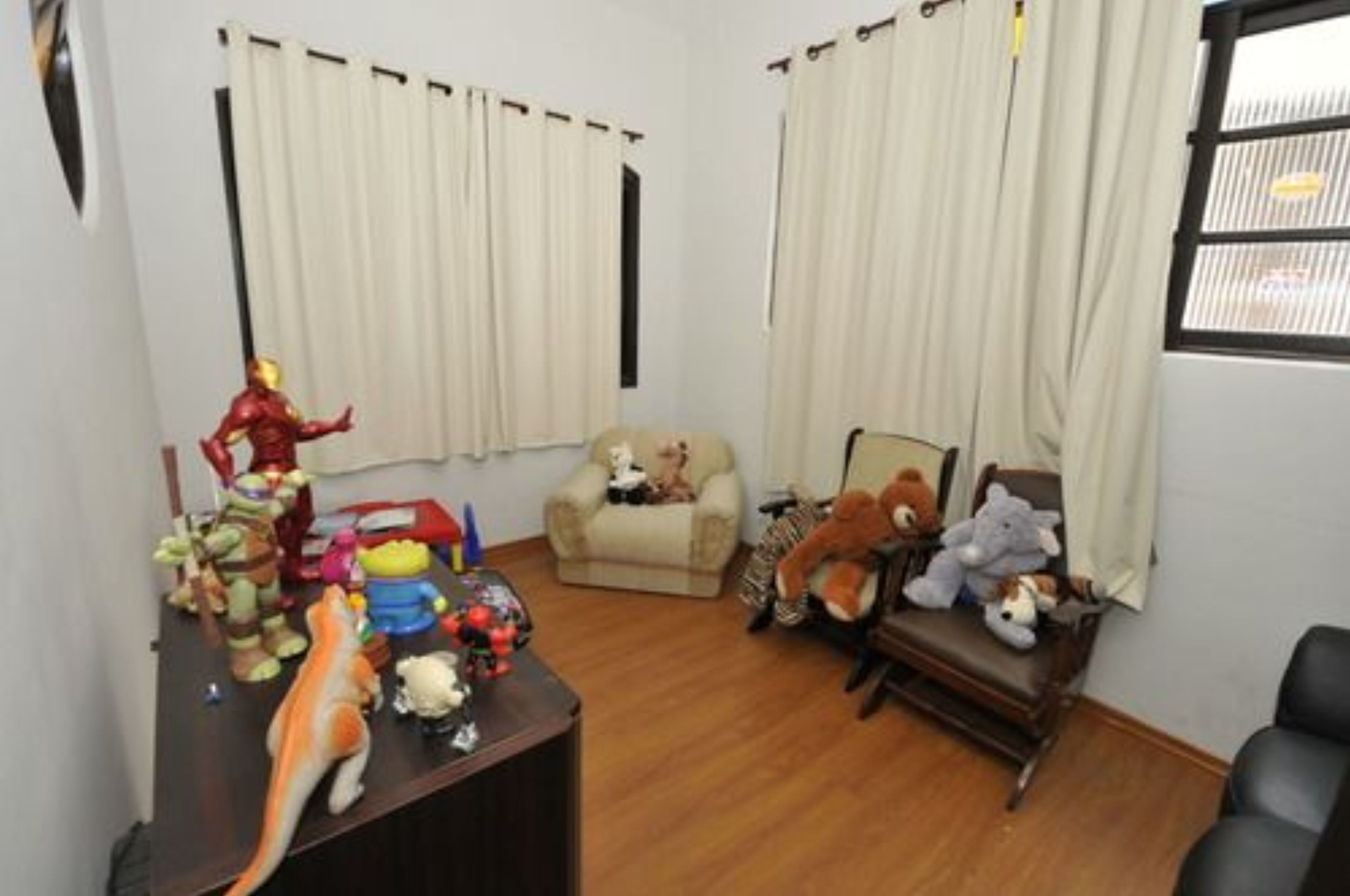 A room with a wooden floor, a desk with various toys, a beige armchair, and a window with curtains.
