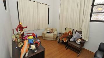 A room with a wooden floor, a desk with various toys, a beige armchair, and a window with curtains.