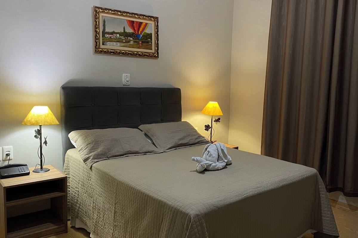 Una cama bien tendida con una manta gris, dos lámparas de noche, un cuadro en la pared y una ventana con cortinas.