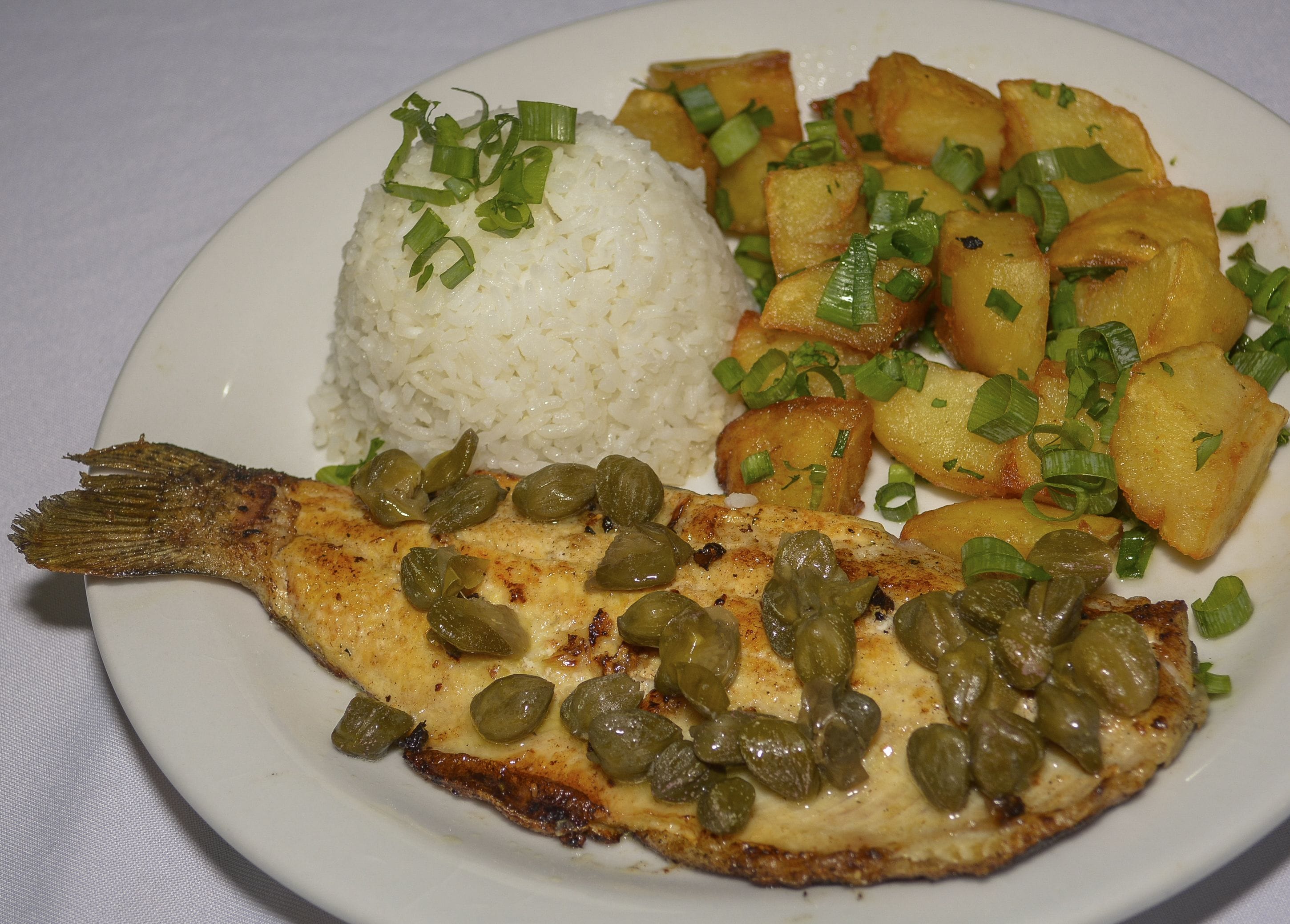 A plate with rice, fried plantains, and a fish topped with capers.