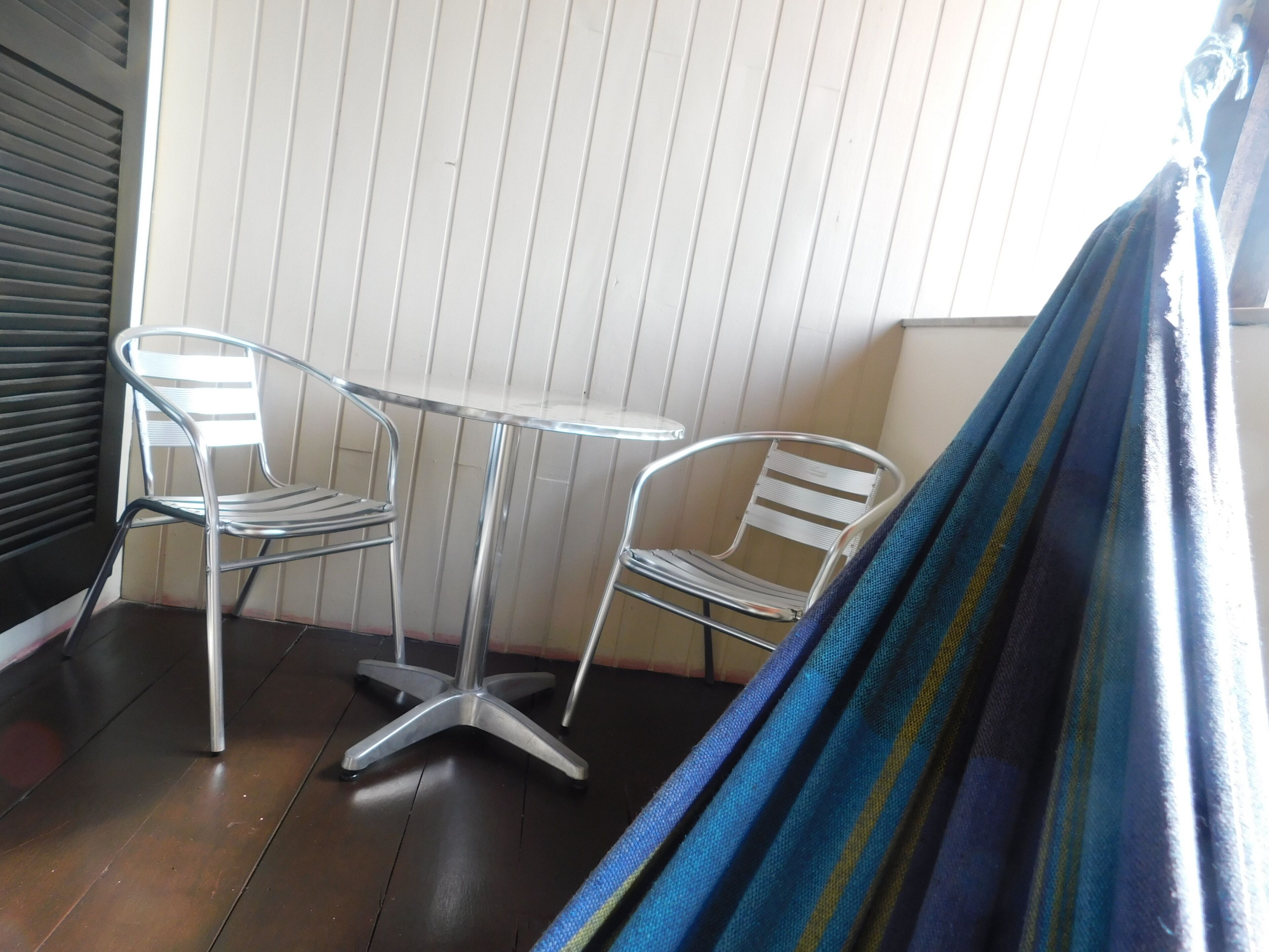A corner room with a metal-framed table and two chairs, a dark wooden floor, and a striped fabric draped over a window.