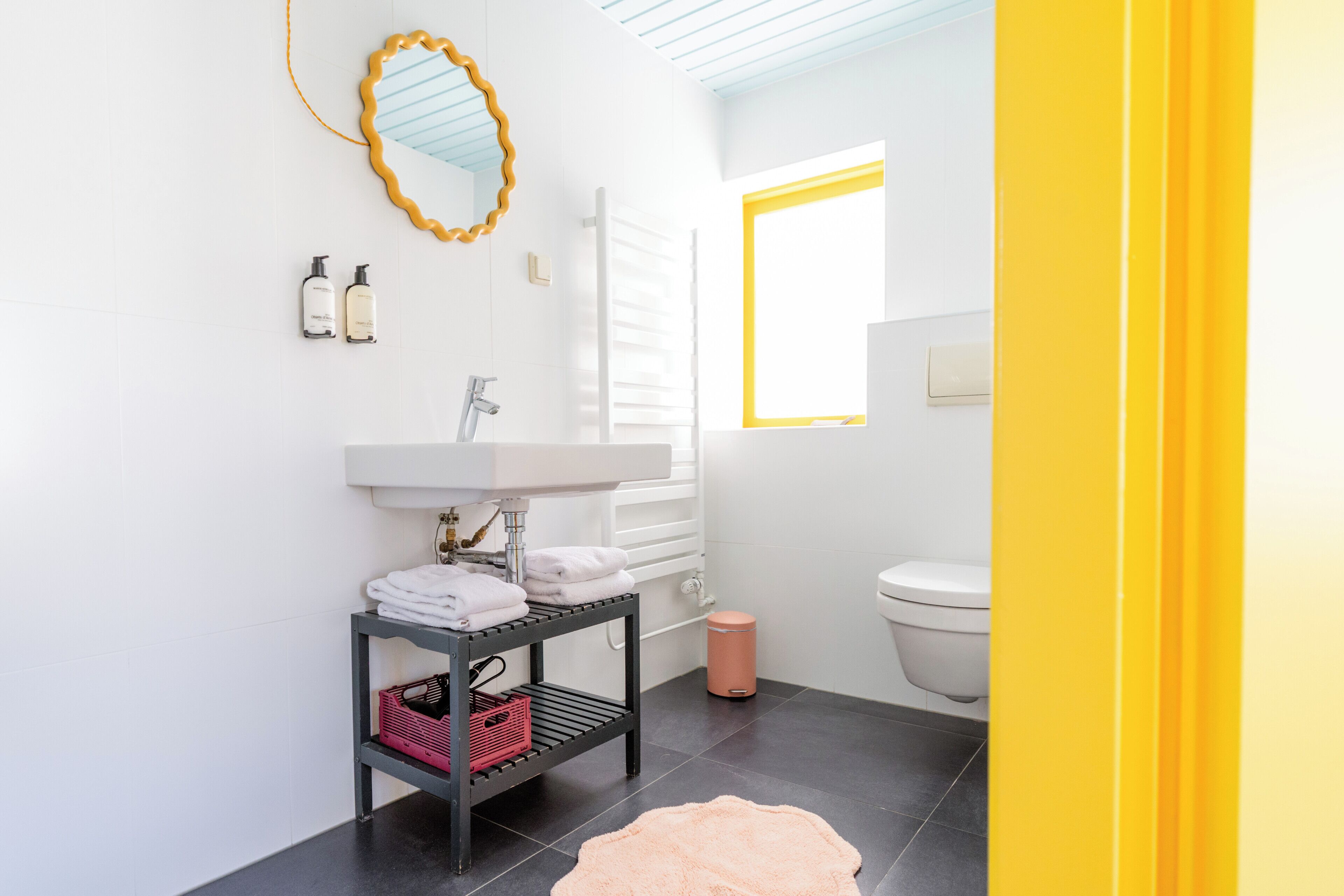 Un bagno moderno con un lavandino bianco, uno specchio con cornice gialla, uno scaffale nero con asciugamani e un tappeto rosa.