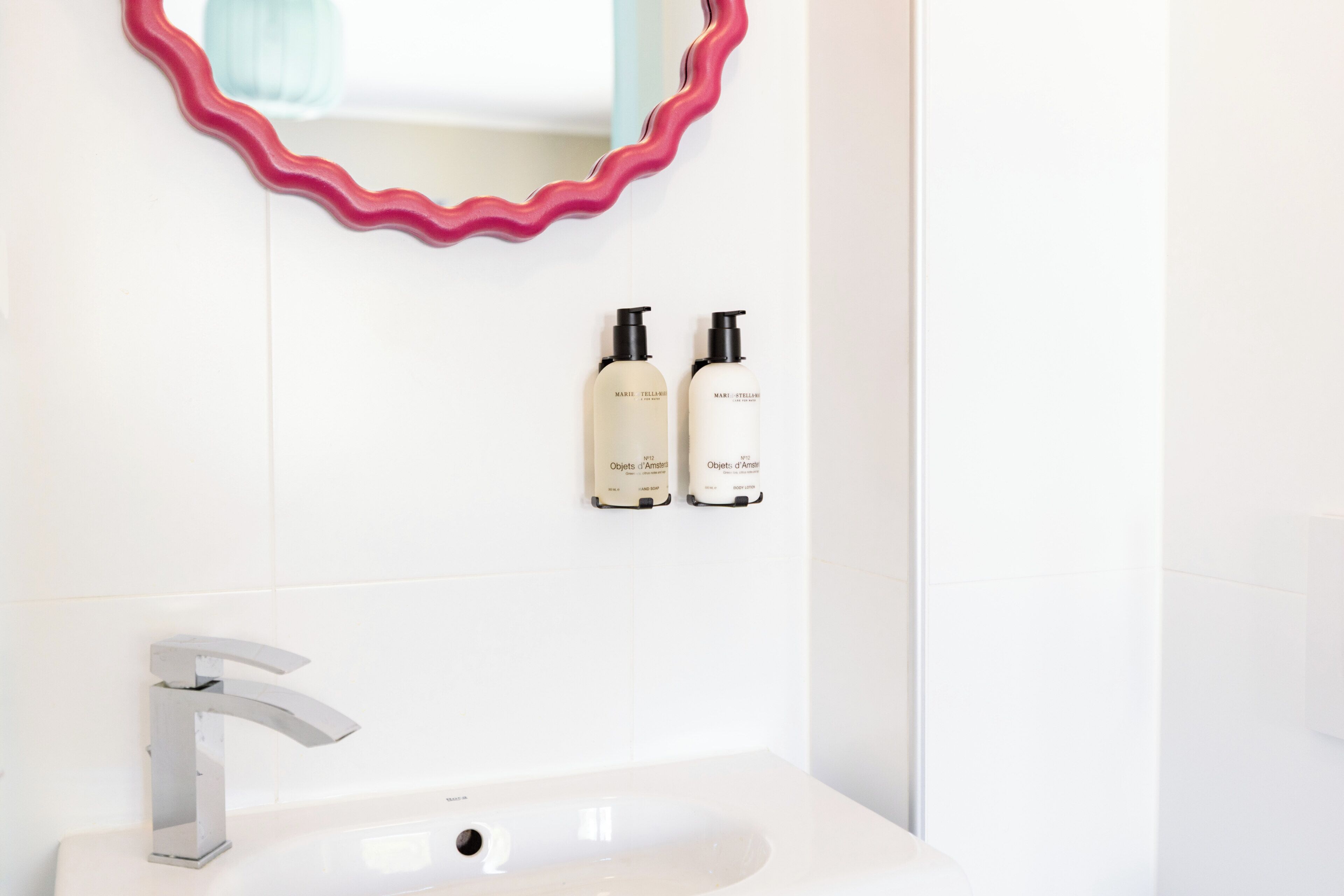 Un baño con un lavabo blanco, un grifo plateado, un espejo con marco rosa y dos botellas de productos para el cuidado de la piel en la pared.