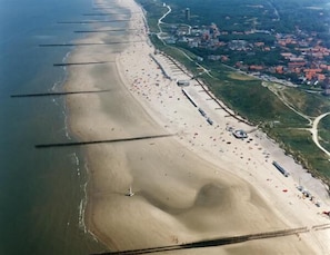Vista aérea de uma praia com uma extensa faixa de areia, algumas embarcações e uma pequena cidade ao fundo.
