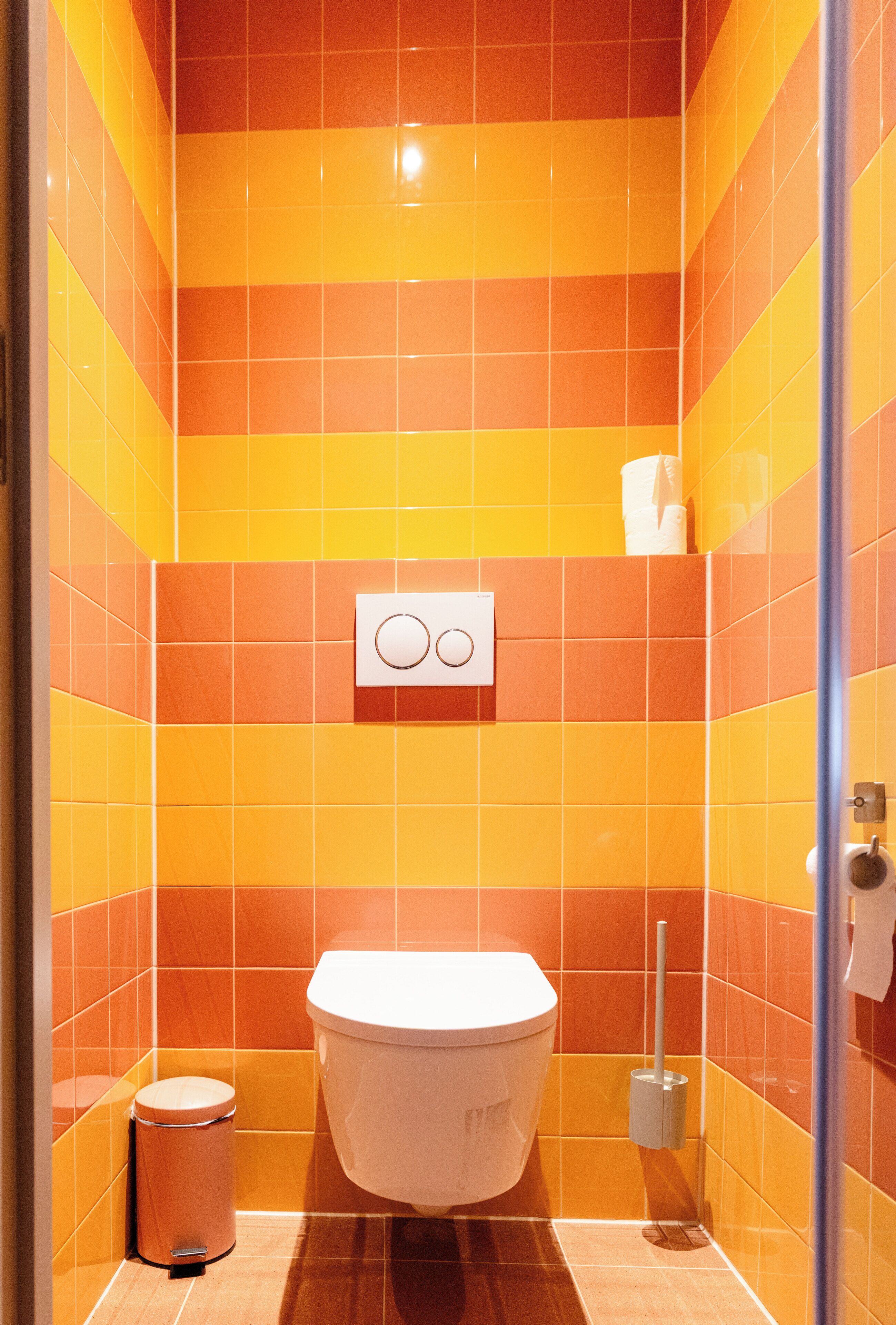 A bathroom with orange tiles, a white toilet, a trash bin, and a paper towel dispenser.