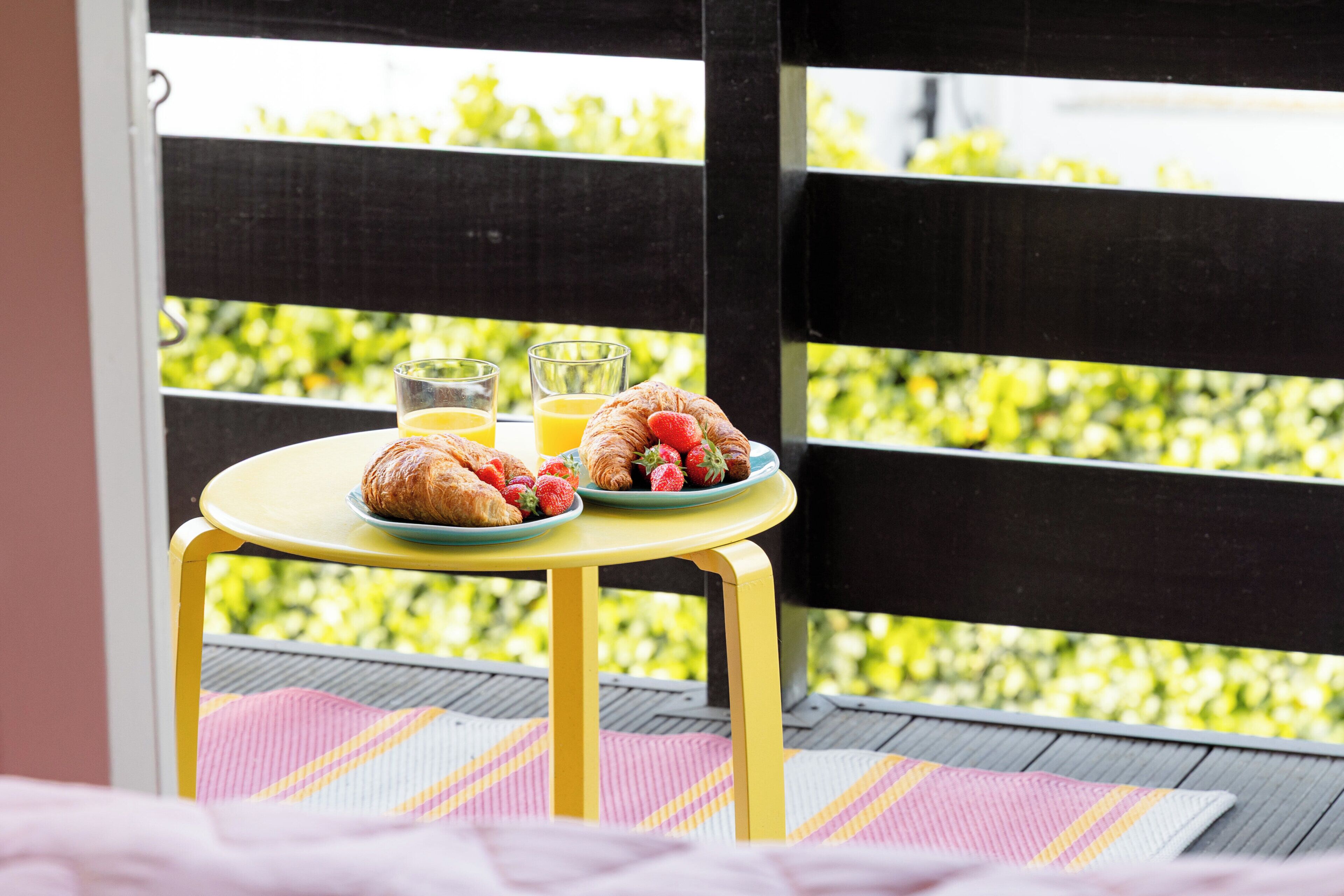Une table jaune dressée pour le petit-déjeuner, avec des croissants, des fraises et du jus d’orange, et une vue sur la verdure à travers une clôture noire.