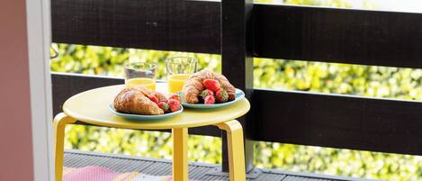 Une table jaune dressée pour le petit-déjeuner, avec des croissants, des fraises et du jus d’orange, et une vue sur la verdure à travers une clôture noire.