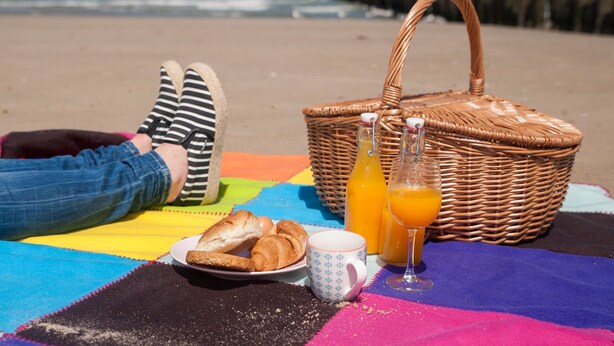 A beach picnic setup with a basket, croissants, a mug, and orange juice on a colorful blanket. - Domburg4you (Domburg)