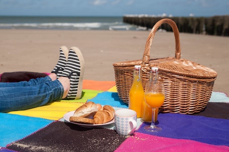 A beach picnic setup with a basket, croissants, a mug, and orange juice on a colorful blanket.