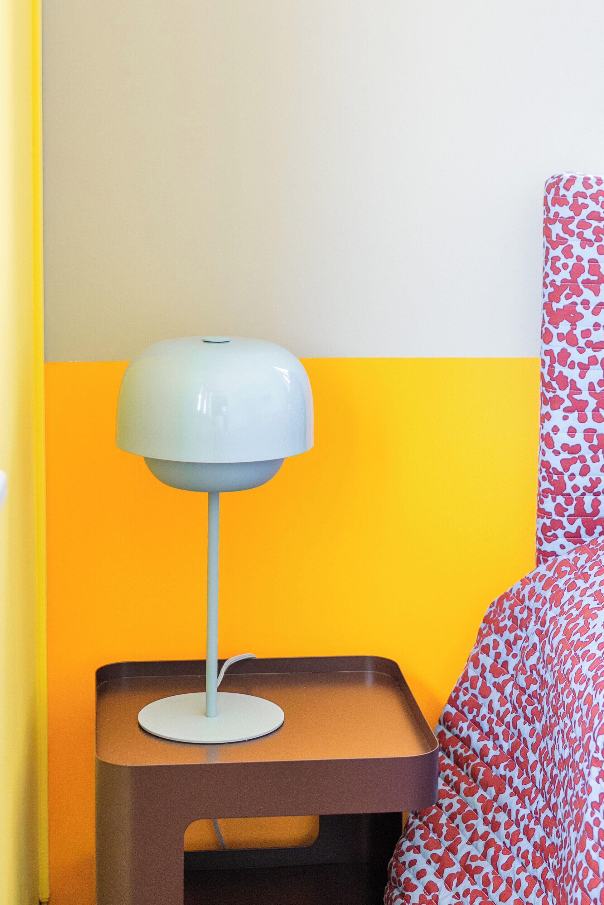 Una cama con cabecera estampada, una mesita de noche con lámpara y una pared con diseño en dos tonos.