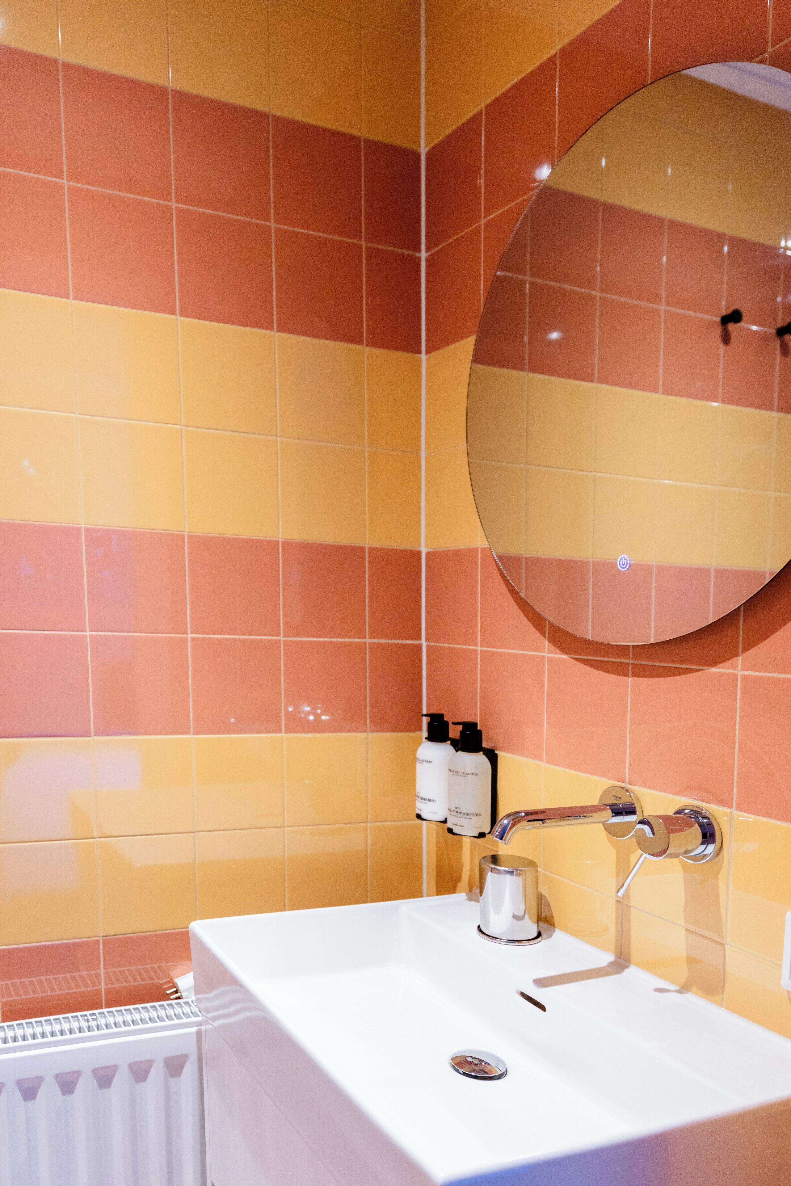 Een badkamer met oranje en gele betegelde wanden, een ronde spiegel, een wit wastafelmeubel en een handdoekrek.