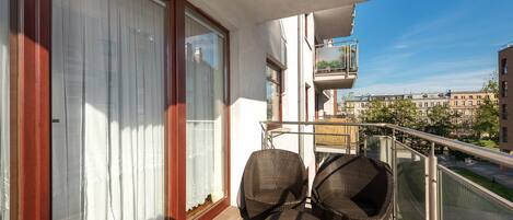 Un balcón con mobiliario de mimbre, barandilla de cristal y vistas a edificios al fondo.