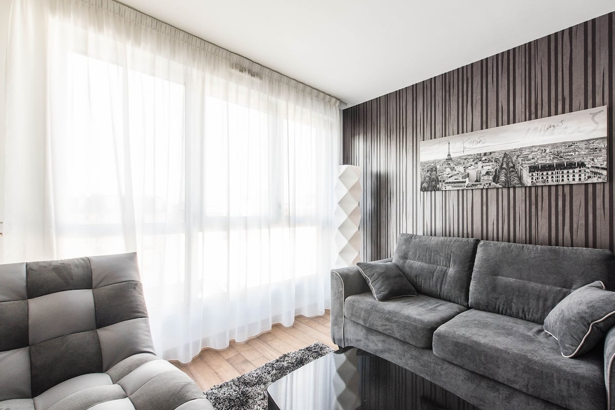 Un salon avec un canapé gris, un fauteuil gris et une œuvre d’art représentant un paysage urbain en noir et blanc accrochée au mur.