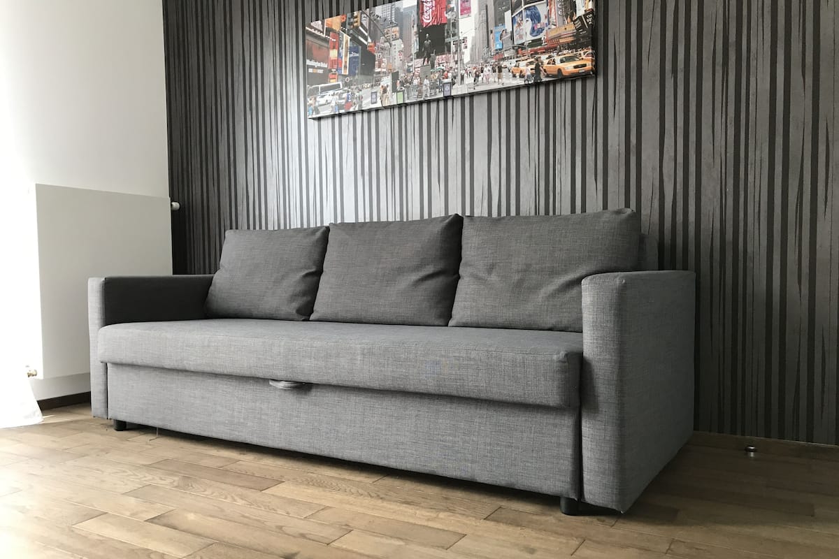 Un canapé gris avec deux coussins, dans une pièce dont le sol est en bois et un des murs est rayé.