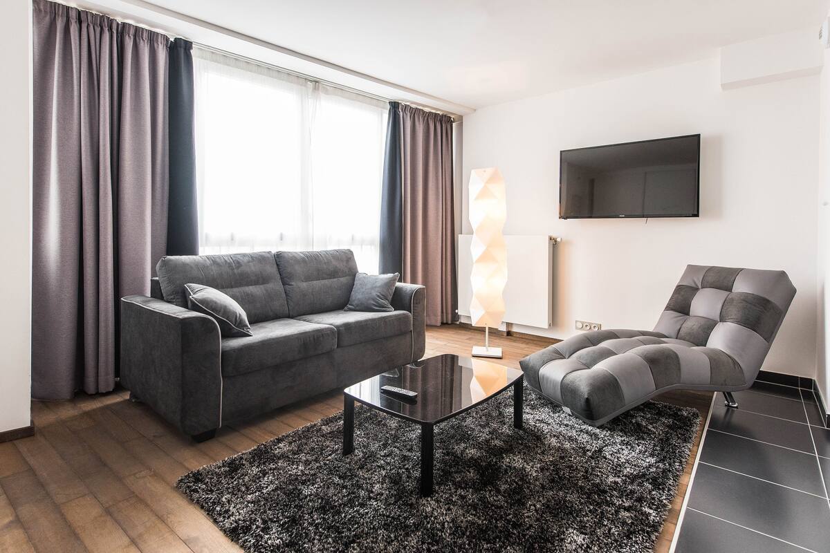 Un salon moderne avec un canapé gris, une table basse noire et un fauteuil gris. Un téléviseur à écran plat est fixé au mur.