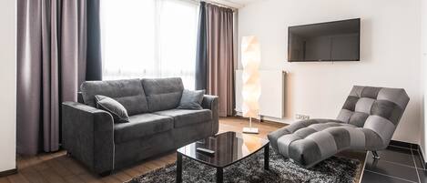 A modern living room with a grey sofa, a black coffee table, and a grey armchair. A flat-screen TV is mounted on the wall.