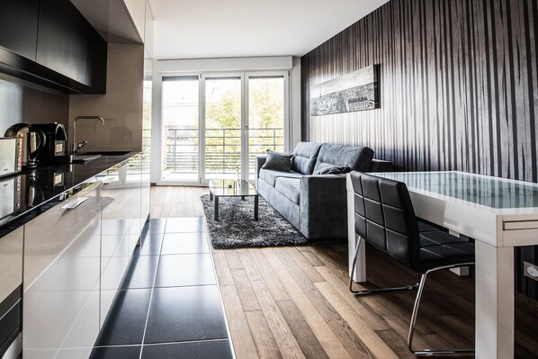 A modern kitchen with a black countertop, a built-in oven, and a coffee machine. There is a grey sofa with a black rug in front of it, and a dining table with chairs.