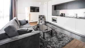 A modern living room with a grey sofa, a black coffee table, and a black and white rug. There is a kitchen area with white cabinets and a flat-screen TV mounted on the wall. - Les Appartements Paris Clichy (Clichy)